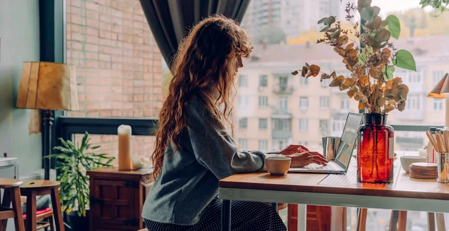 A woman working remotely on a laptop by a window with a city view — comparing the D7 and D8 visas for Portugal